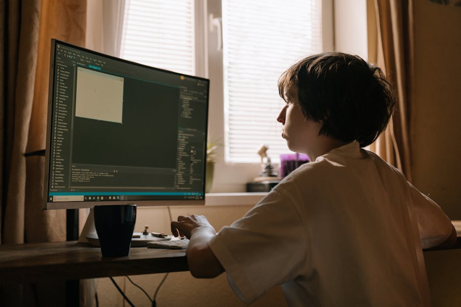 A person with short, dark hair is seated at a wooden desk in a room with natural light coming through two windows with white blinds. They are wearing a white shirt and are focused on a large computer